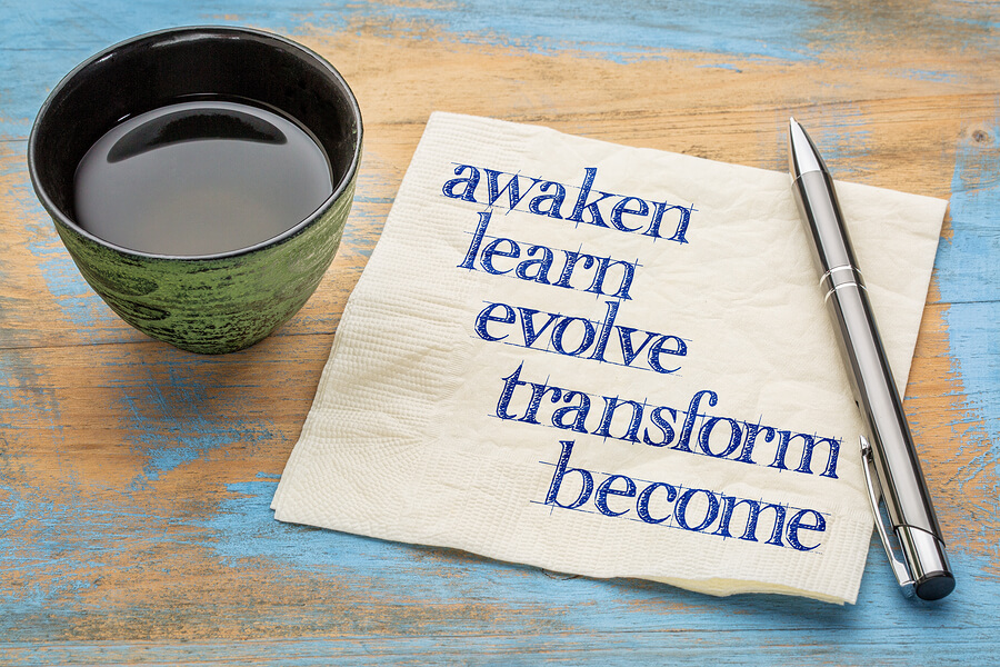 A cup of coffee with the words awaken, learn, evolve, transform, & become on a napkin